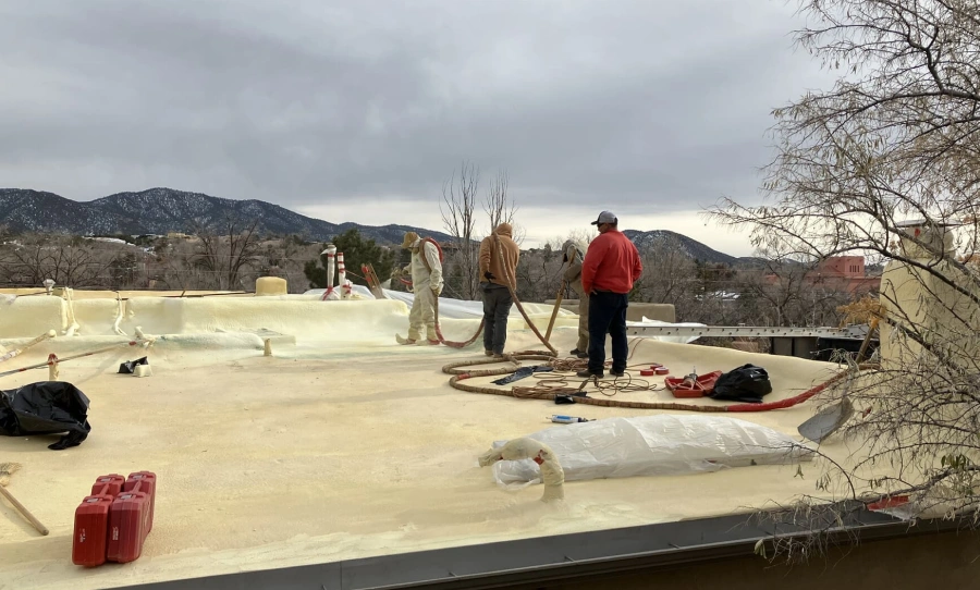 Four workers on a light yellow foam roof with dark mountains in the background under a cloudy sky.